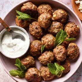 A bowl of perfectly browned lamb meatballs garnished with fresh mint, served with a side of creamy tzatziki.