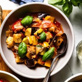 A bowl of rustic stewed vegetables, including eggplant and peppers, topped with fresh basil.